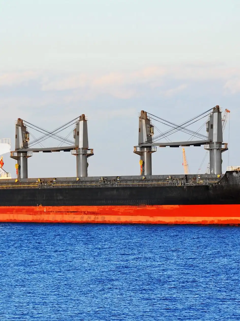 Black-hulled bulk carrier with red boot-top and four deck cranes riding at anchor on calm water