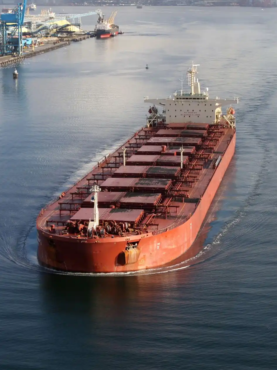 Charter a bulk carrier: red-hulled vessel approaching port with hatch covers open, aerial daylight view