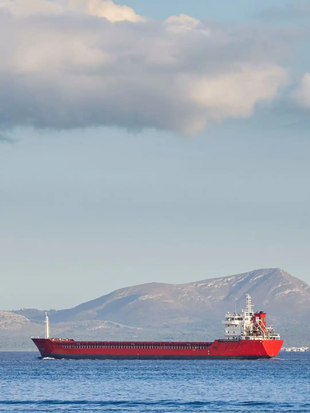 A red bulk carrier underway on calm water with a mountainous coastline behind