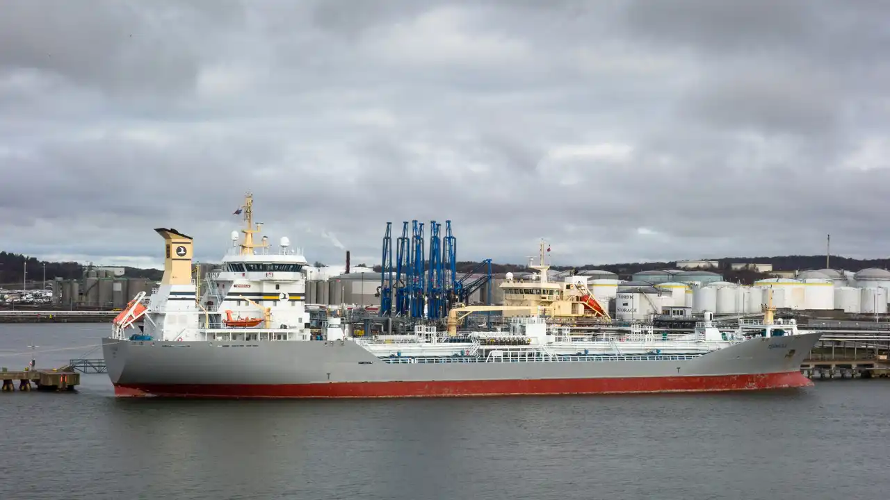 A stainless-tank chemical tanker berthed at a liquid-bulk terminal, with shore storage tanks and loading gantries behind