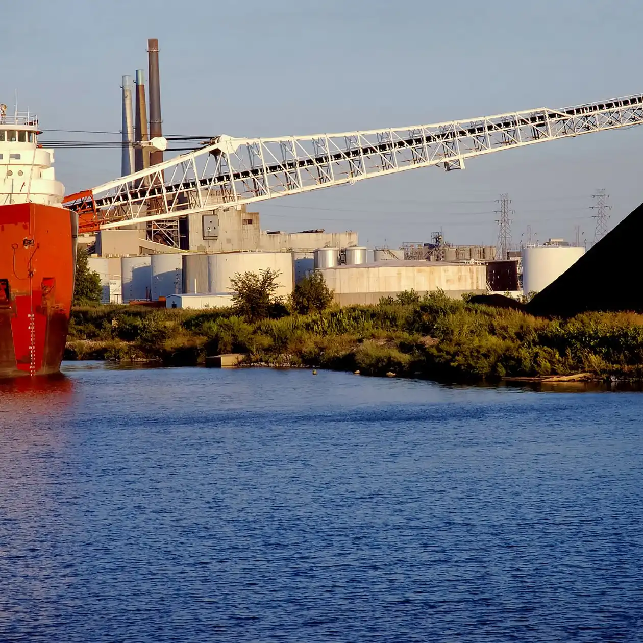 A red bulk carrier at an industrial berth beside a dark bulk pile and conveyor