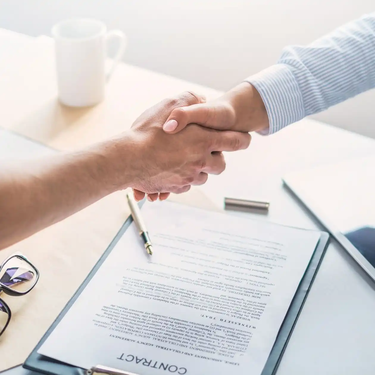 Two people shaking hands over a desk with contract documents and a pen