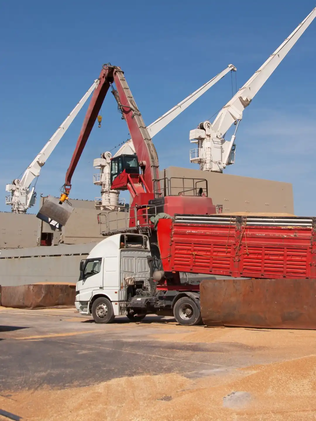 A crane loading grain onto a cargo ship beside a truck at a terminal
