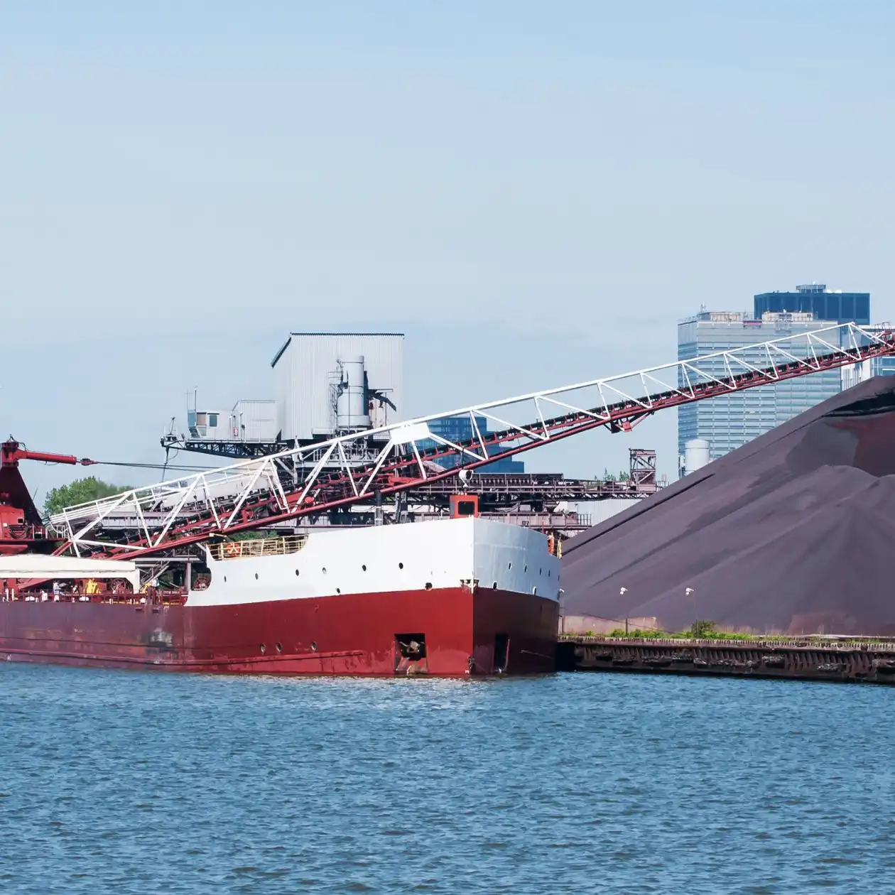 A red bulk carrier at a port handling dark iron ore via a shore conveyor
