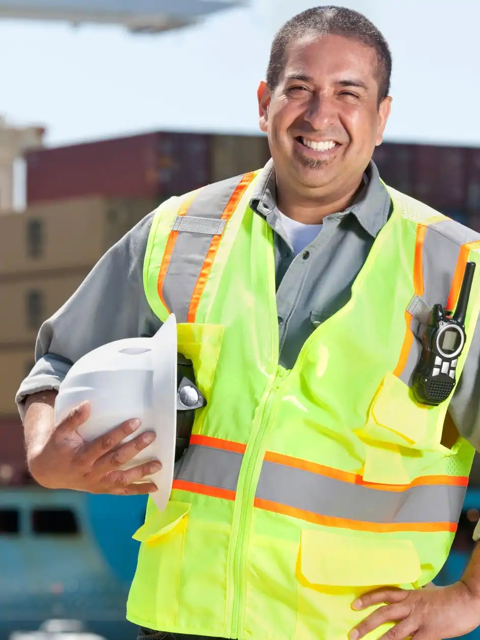 A port worker in a hi-vis vest holding a hard hat and radio, standing in front of a container-laden vessel