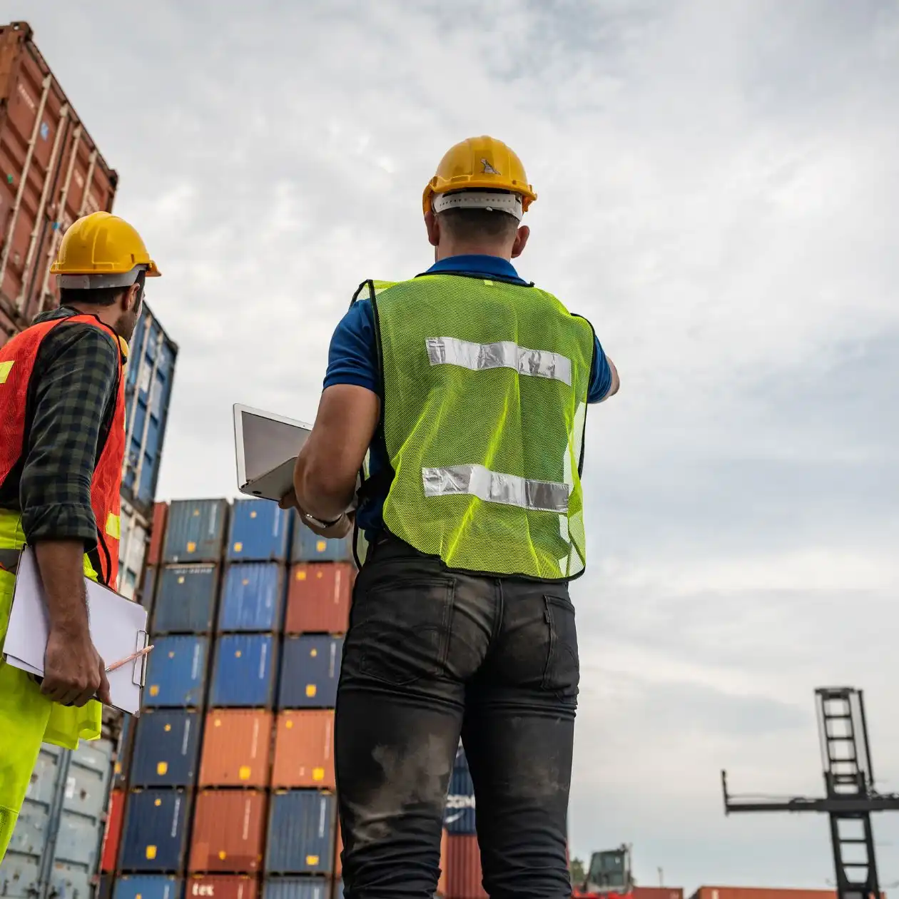 Two port workers in hi-vis vests and hard hats with a clipboard before containers