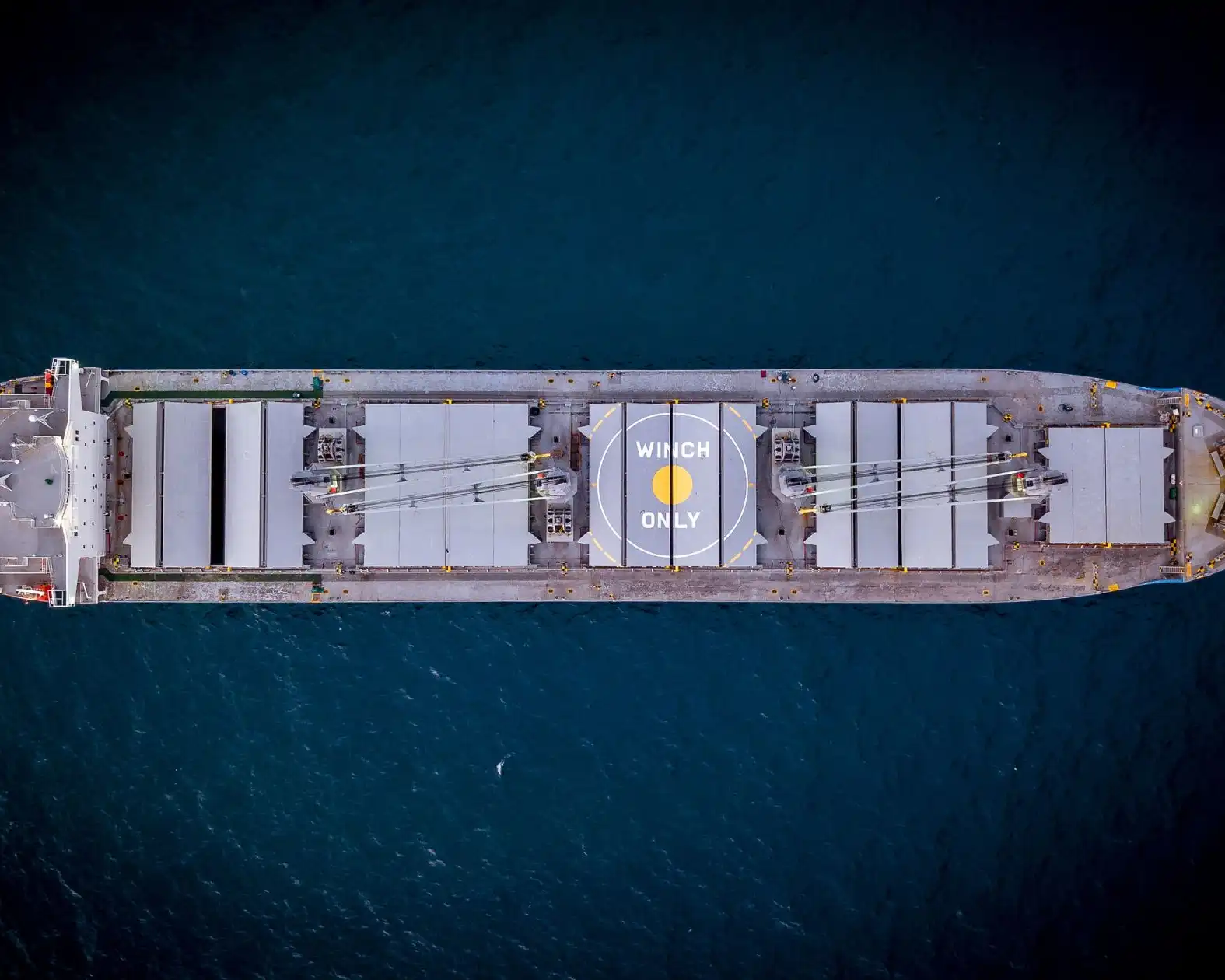 Aerial top-down view of a bulk carrier with hatch covers and deck cranes underway on dark blue water