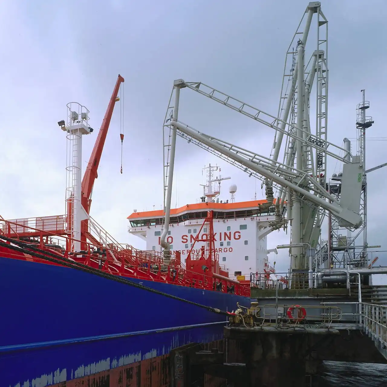A blue-hulled product tanker berthed alongside a terminal, with white marine loading arms connected over its deck