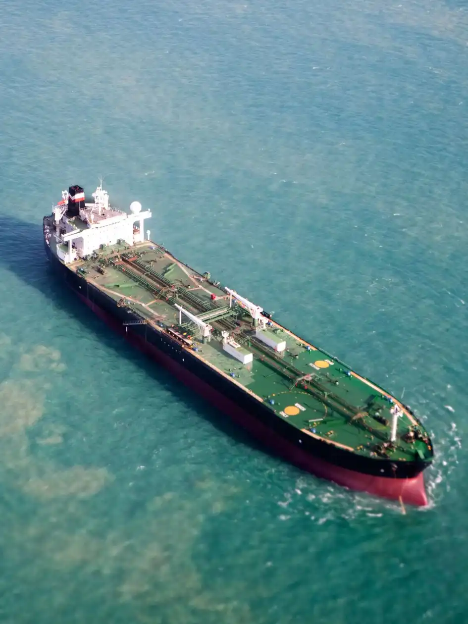 Aerial view of a dark-hulled tanker with a white superstructure underway through greenish-blue sea, deck pipework visible