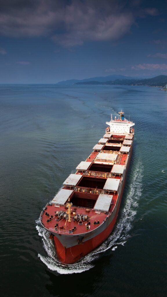 A large red cargo ship with white structures on its deck sails through calm waters. The vessel leaves a trail in the water as it moves forward. The horizon shows mountainous terrain under a partly cloudy sky.