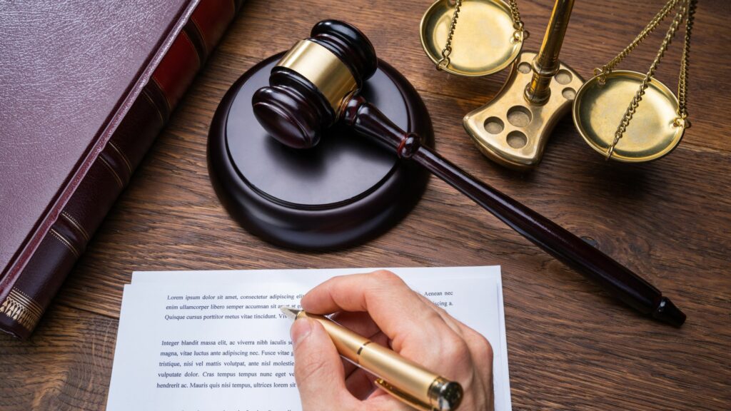 A hand holding a pen is writing on a document placed on a wooden desk. Next to the document are a gavel on a sound block, a pair of golden scales, and a legal book. The scene depicts a law-related setting, perhaps serving as an essential guide to Admiralty Law and Maritime Law.