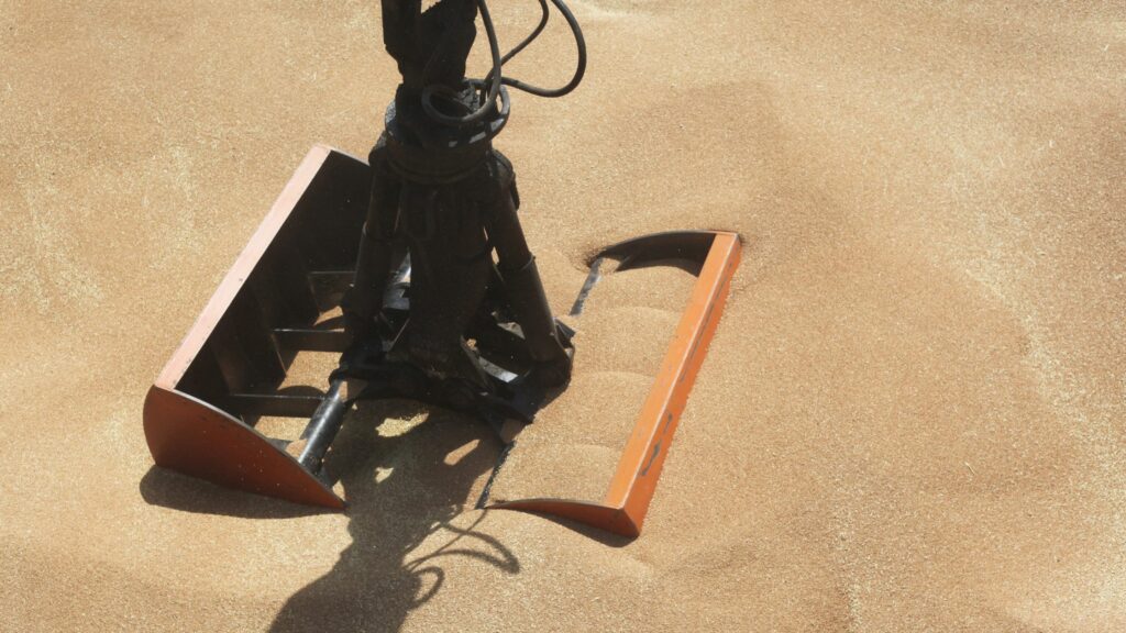 A close-up view of an industrial grabber attached to machinery, partially buried in a pile of golden brown sandy material. The grabber has two large metal orange claws, with shadows cast on the sand indicating sunlight.