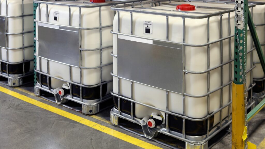Several large industrial containers filled with liquid are arranged in a warehouse. The containers are made of white plastic and are encased in metal cages. Each container has a red cap on top and a valve at the bottom for dispensing the liquid.
