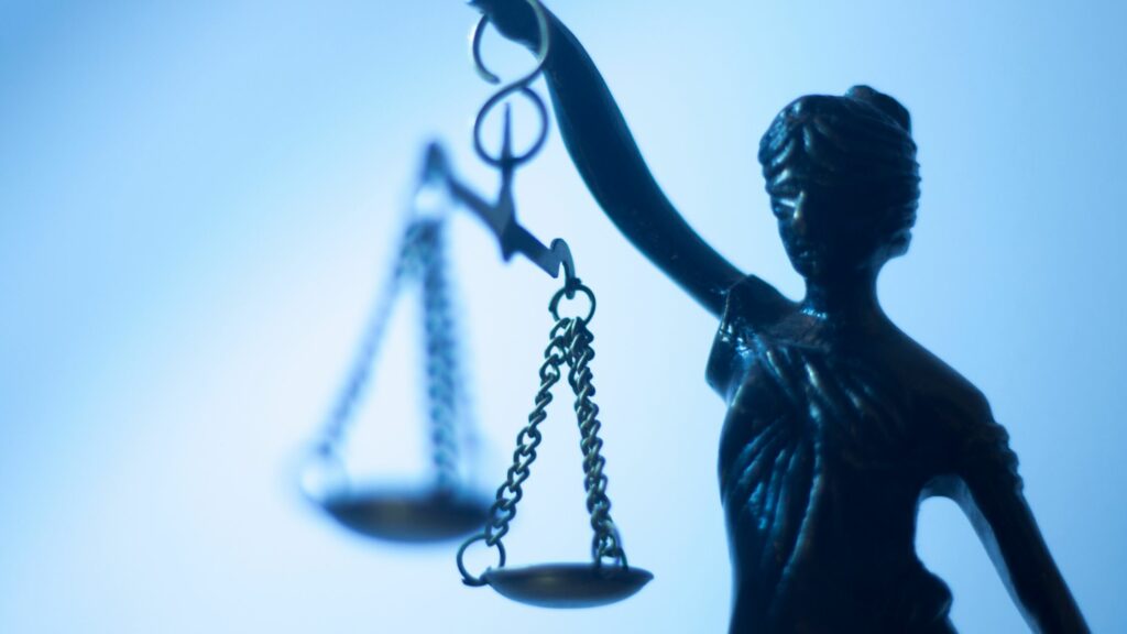 Close-up of a bronze statue of Lady Justice holding scales, reminiscent of a guide in maritime law. The background is a gradient of light blue, giving the scene a cool tone. The scales are in sharp focus, symbolizing impartiality in justice. The statue's features are slightly blurred.