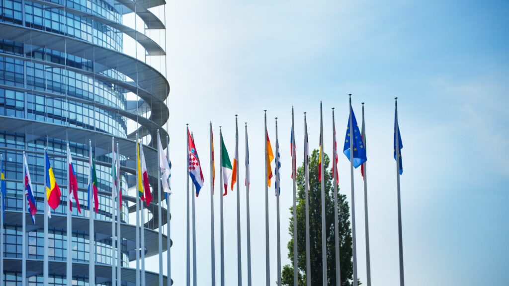 A series of national flags stand in a row on tall flagpoles outside a modern, circular glass building. The blue sky and a tree are visible in the background. The flags represent various countries, including a prominent flag of the European Union, serving as guides for discussions on Maritime Law.